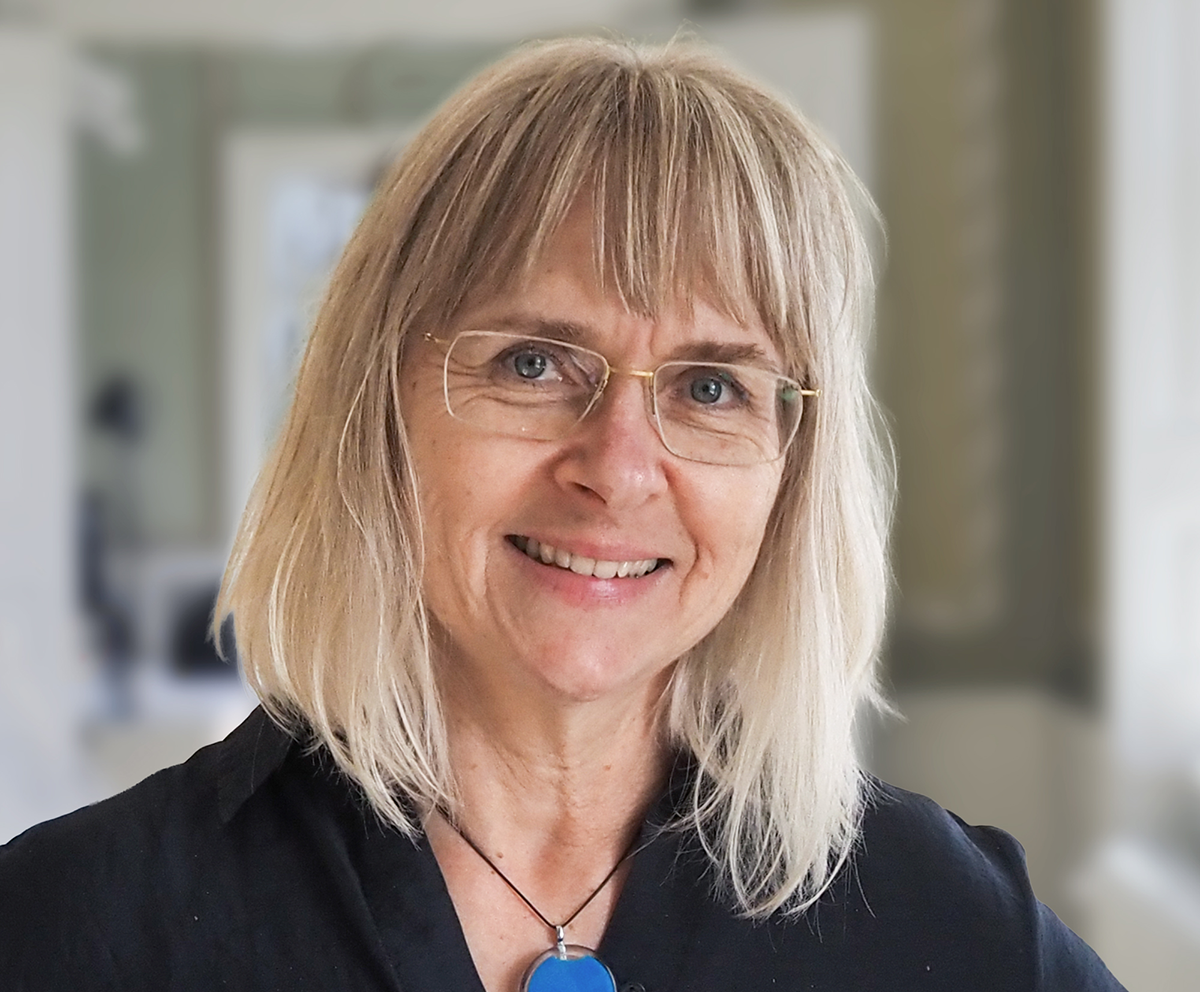 En kvinna i medelåldern med blont hår och glasögon, svart topp och blått hängsmycke ler och står inomhus.