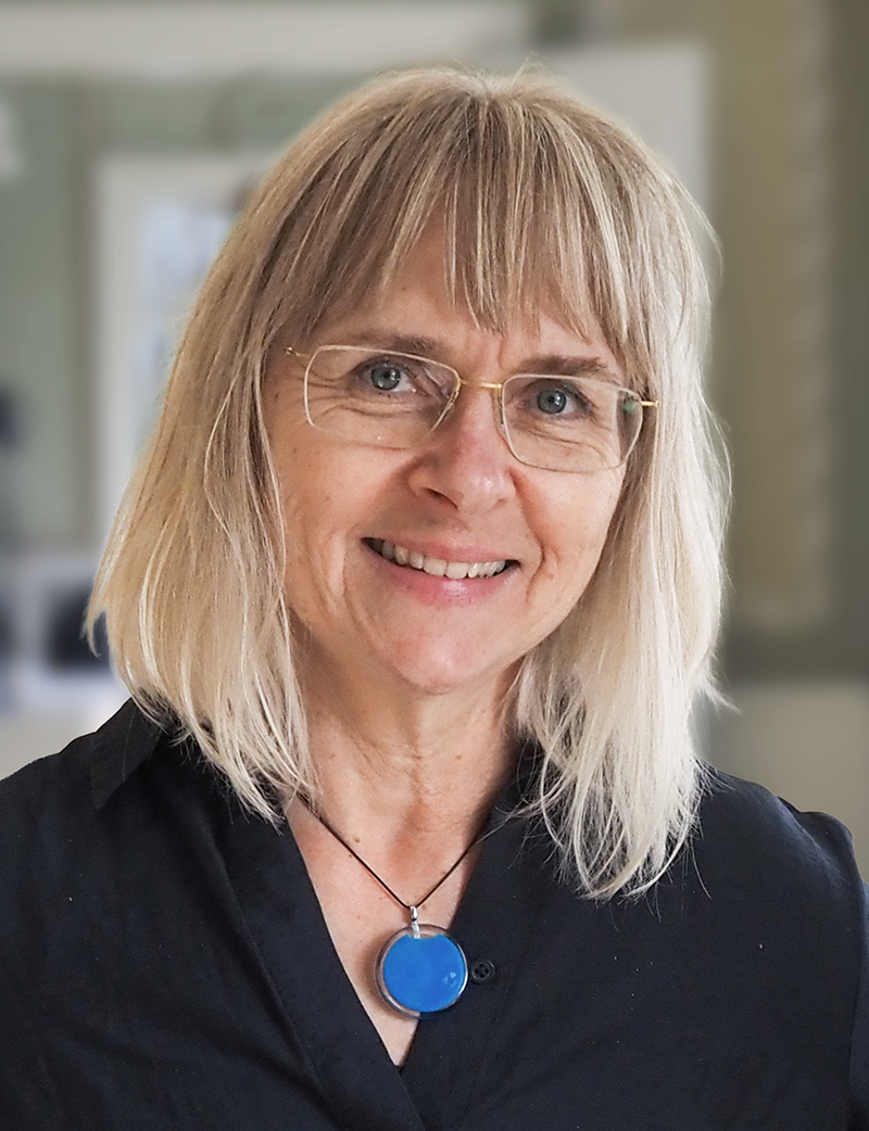 En kvinna med blont hår och glasögon ler mot kameran. Hon har på sig en svart tröja och ett blått hängsmycke. Bakgrunden är suddig.