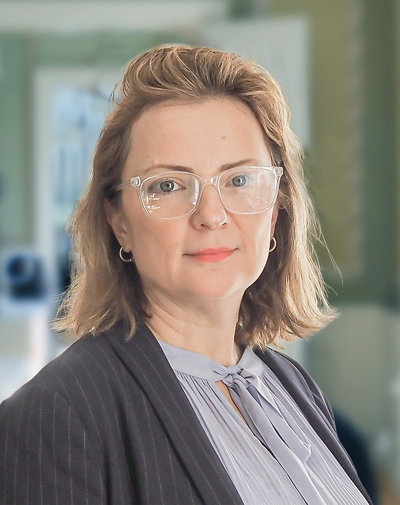 En kvinna med axellångt ljusbrunt hår, bär genomskinliga glasögon, en randig kavaj och en ljusblå blus, står inomhus.