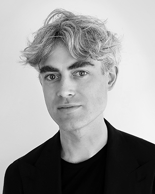 En man med vågigt grått hår har en mörk kavaj och svart skjorta och ser mot kameran, med en ljus bakgrund bakom sig.