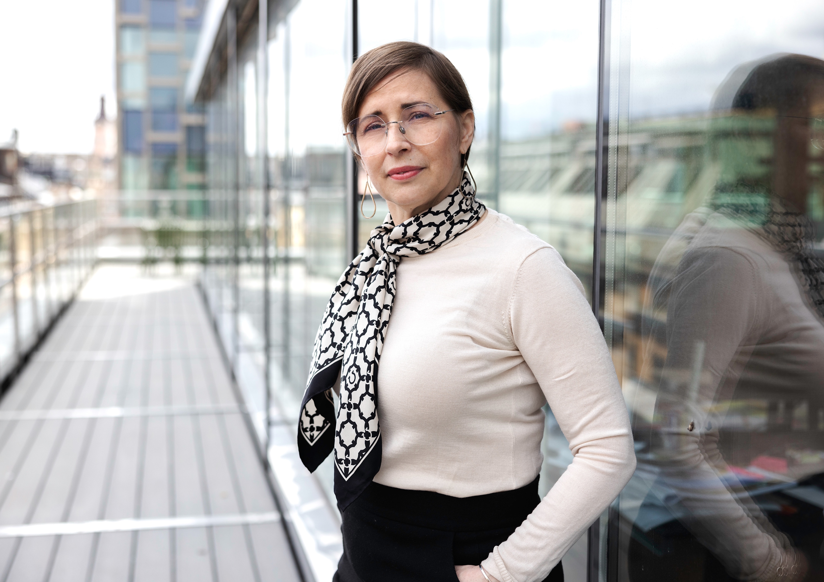 Kvinna med glasögon och mönstrad scarf står på en utomhusgång bredvid en glasvägg och tittar in i kameran.