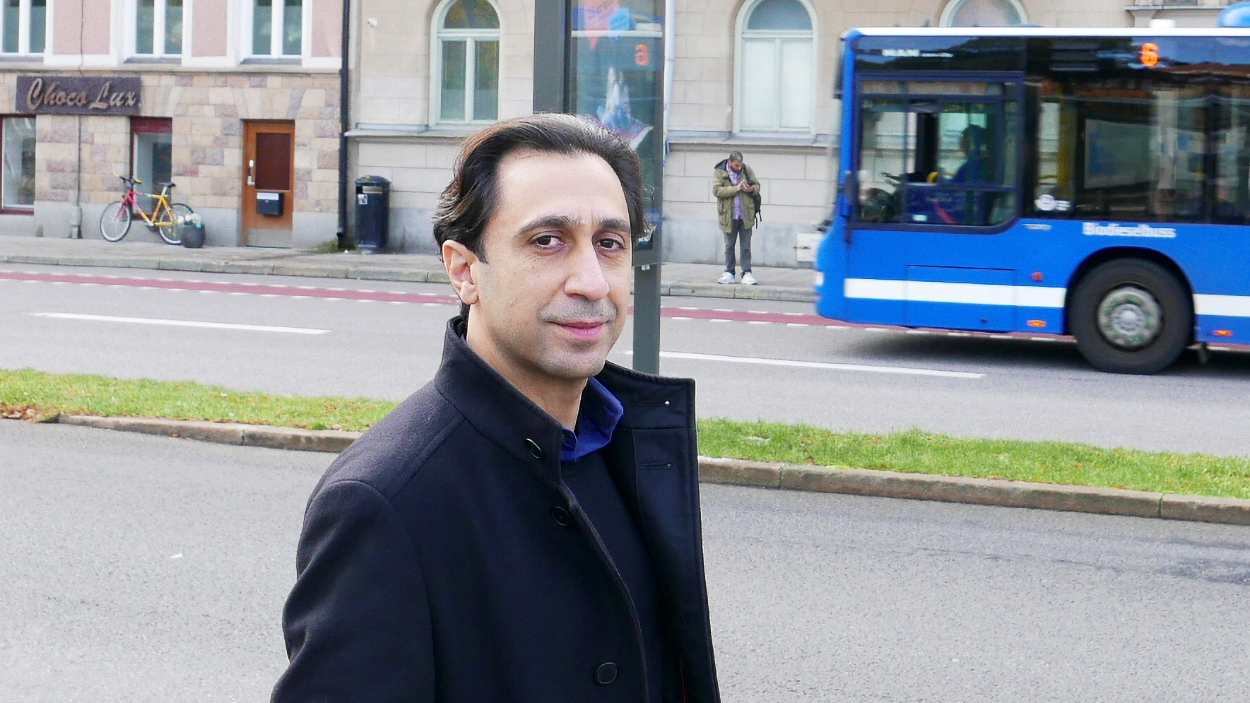 En man med mörk rock står på en stadsgata, en blå buss kör förbi och det finns byggnader i bakgrunden.