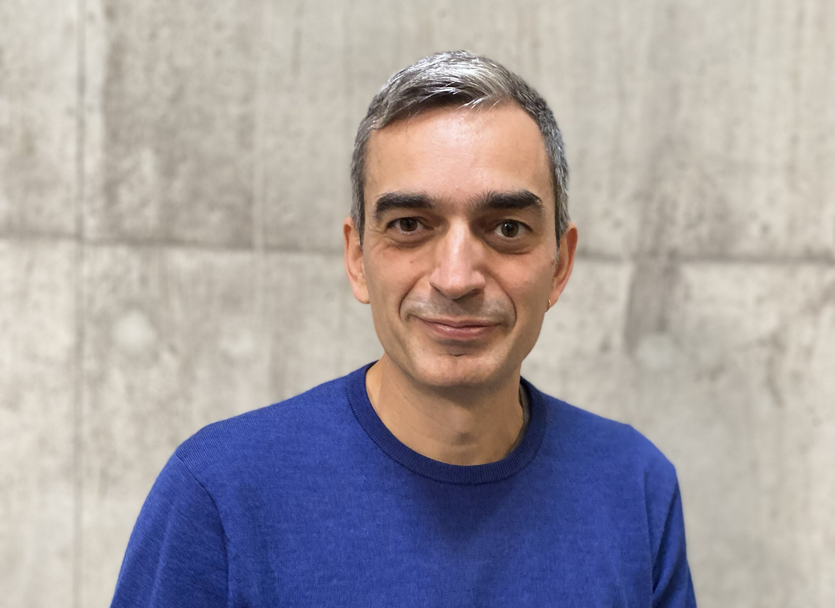 En man med kort grått hår och blå tröja står framför en ljus betongvägg och tittar mot kameran.