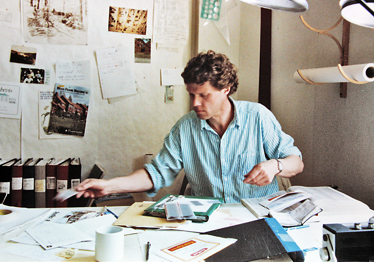 En person sitter vid ett skrivbord och sorterar papper och dokument. På väggen bakom finns böcker, anteckningar och fotografier.