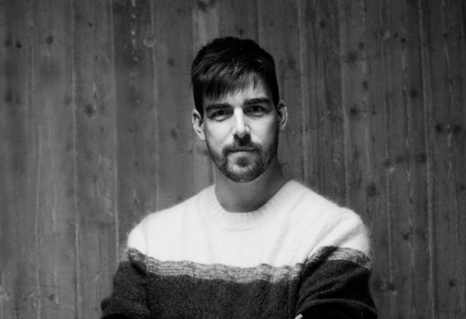 En man med kort mörkt hår och skägg har på sig en randig tröja och står framför en trävägg, vänd mot kameran.