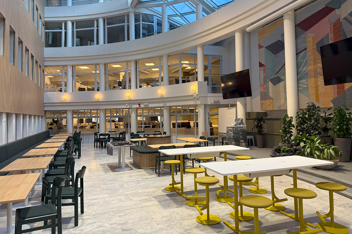 Inomhusatrum med bord, stolar, stora fönster, växter och en geometrisk väggmålning. Flera arbetsytor på övre våningar.