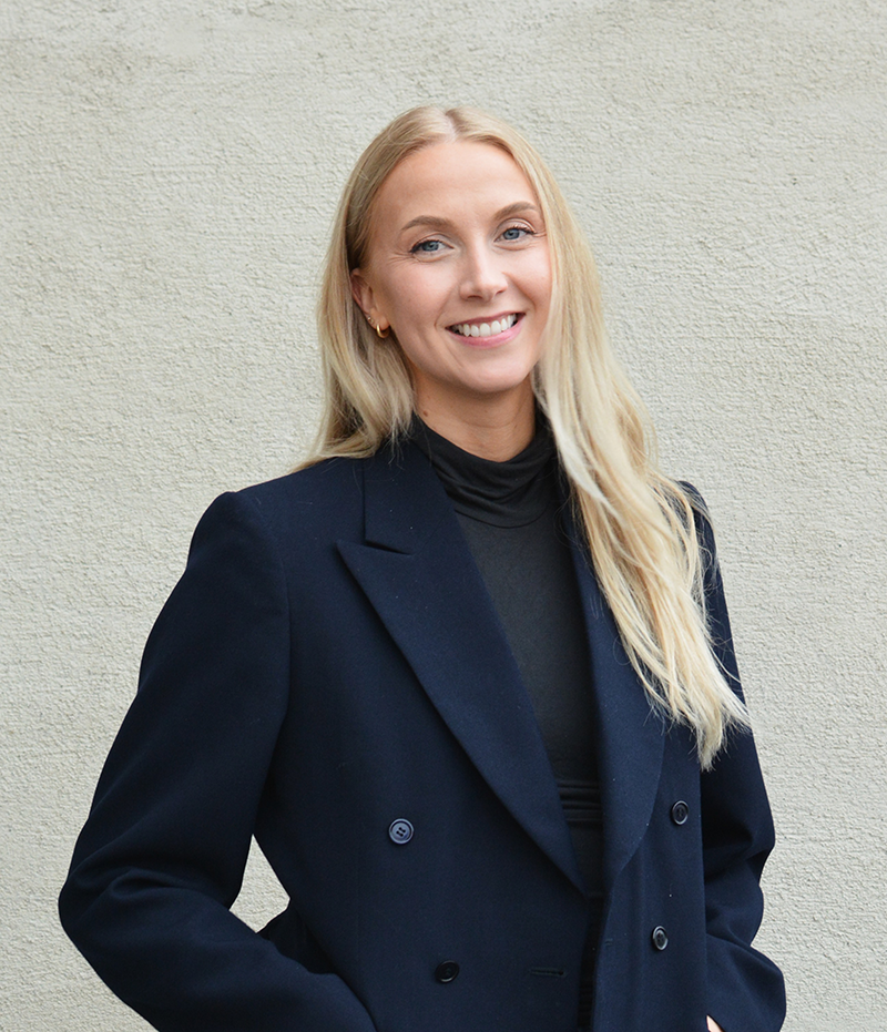 En kvinna med långt blont hår, svart kavaj och svart topp står framför en ljus vägg och ler mot kameran.