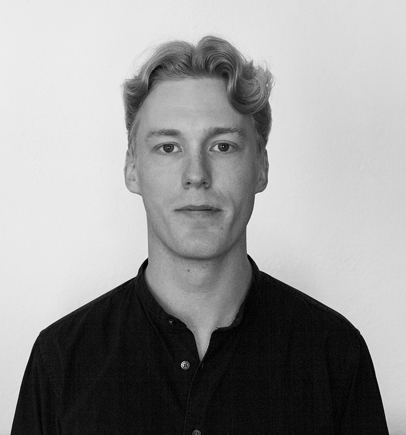 En man med ljust hår och en mörk skjorta står mot en vit bakgrund och tittar rakt mot kameran med neutral min.