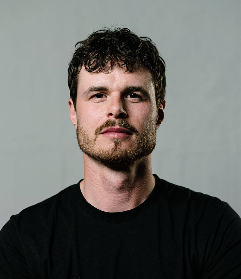 En man med kort mörkt hår, skägg och mustasch har på sig en svart t-shirt och står mot en grå bakgrund.