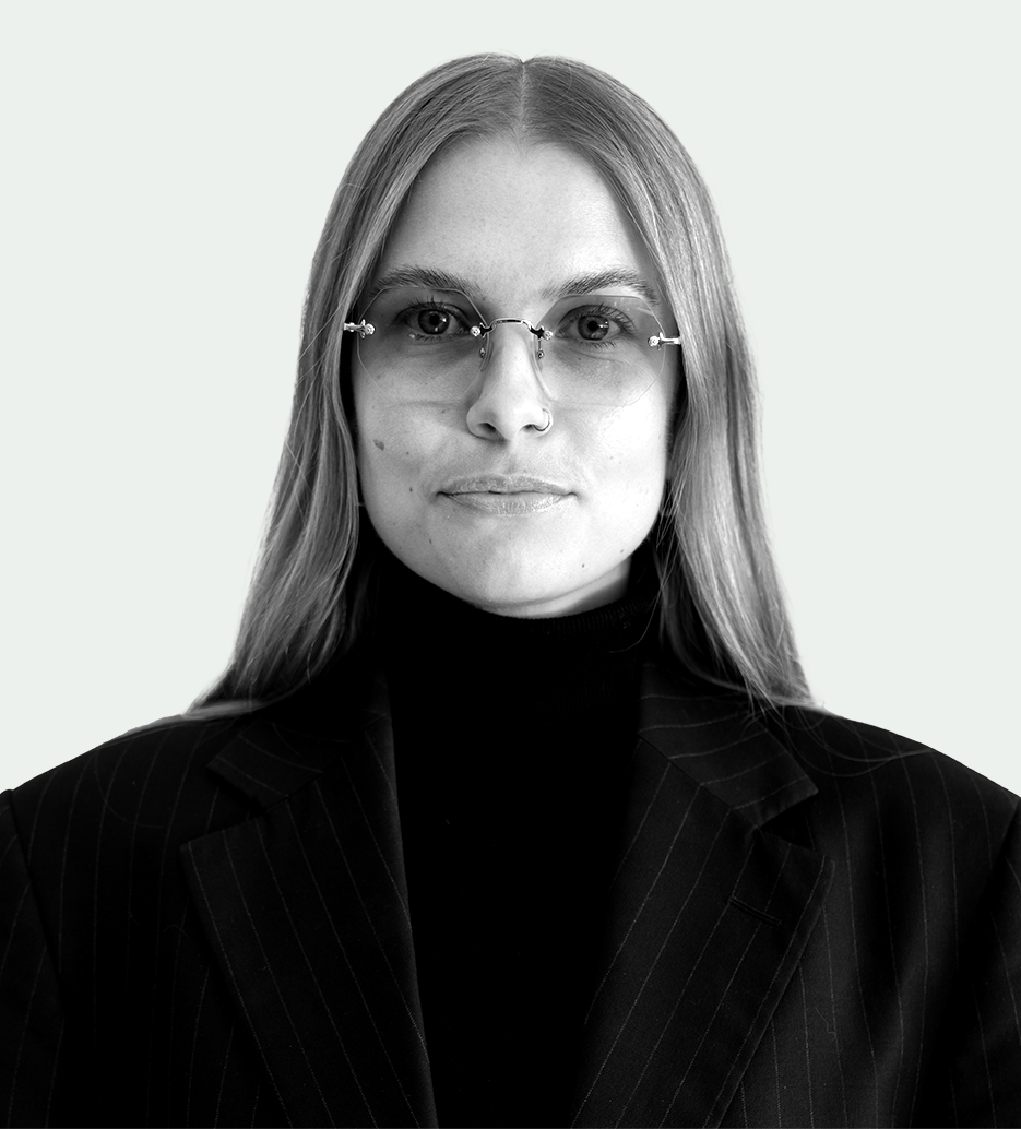 En kvinna med långt, rakt hår har glasögon, en mörk polotröja och kavaj med ränder, vänd framåt mot ljus bakgrund.