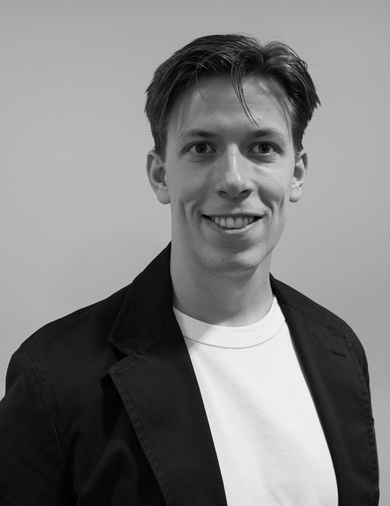 En man med kort hår har på sig en mörk kavaj över en ljus skjorta och ler mot kameran framför en enkel bakgrund.