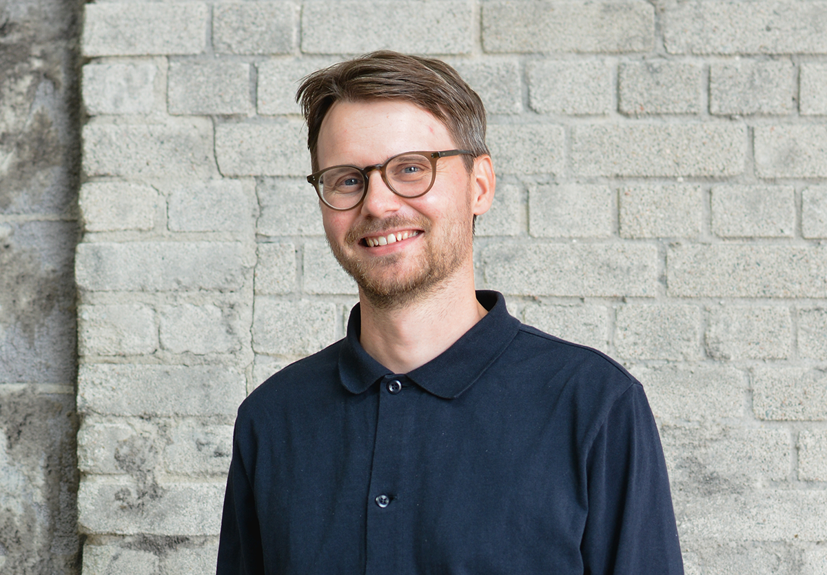 En man med kort brunt hår och glasögon står framför en ljusgrå tegelvägg. Han har på sig en mörk skjorta med knappar.