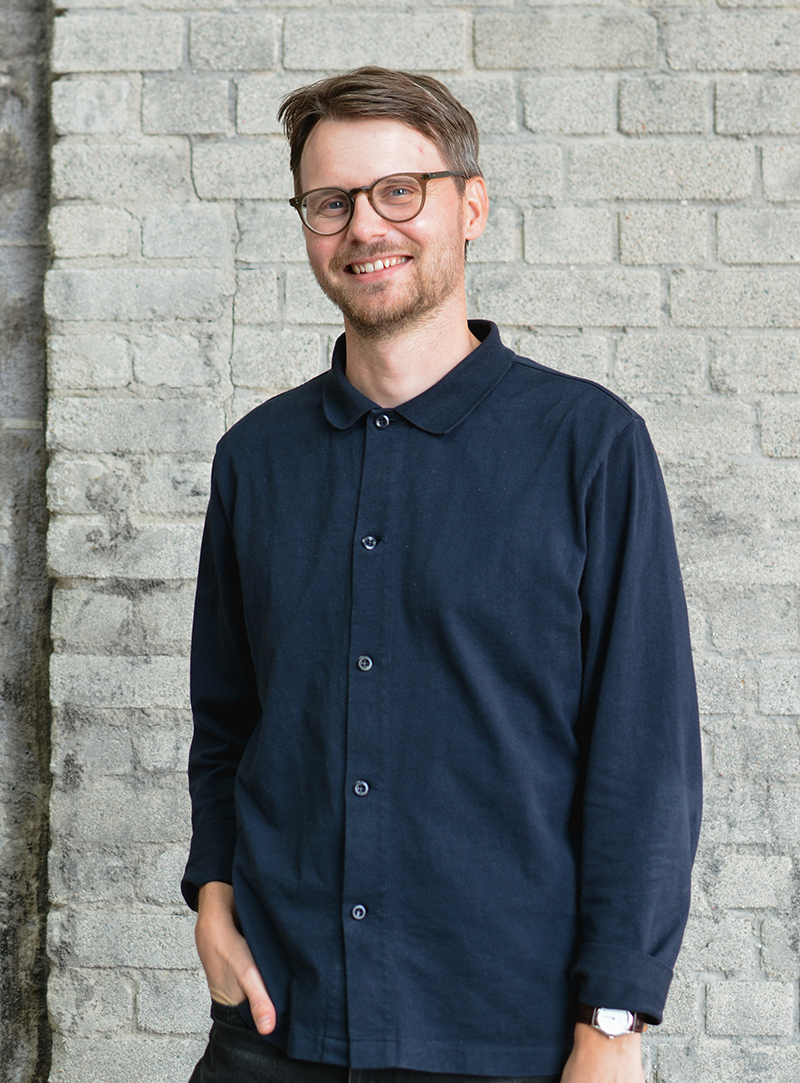 En man med glasögon och kort hår står framför en ljusgrå tegelvägg, klädd i en mörkblå skjorta med ena handen i fickan.