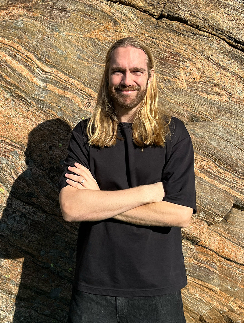 En man med långt blont hår står utomhus mot en stenmur, med armarna i kors, iförd svart t-shirt och jeans.