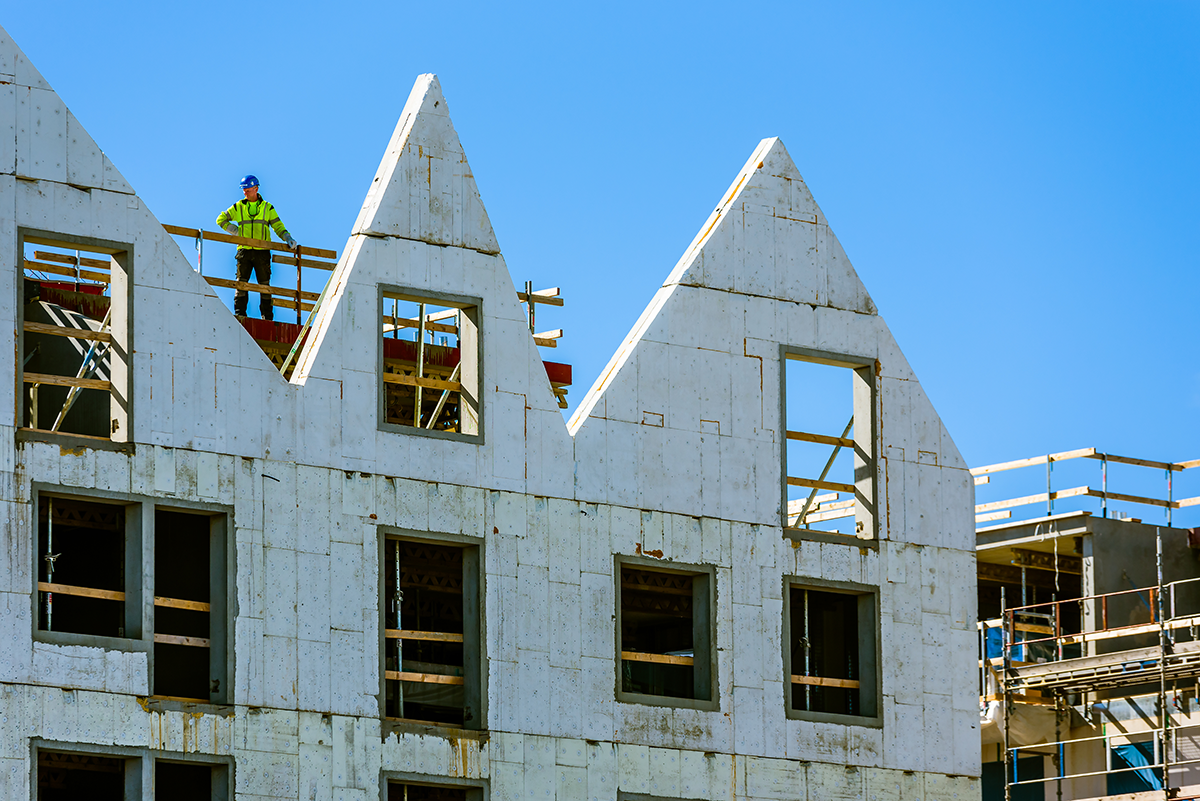 En byggarbetare med skyddsutrustning står på översta våningen av en delvis färdig byggnad med spetsiga tak mot blå himmel.