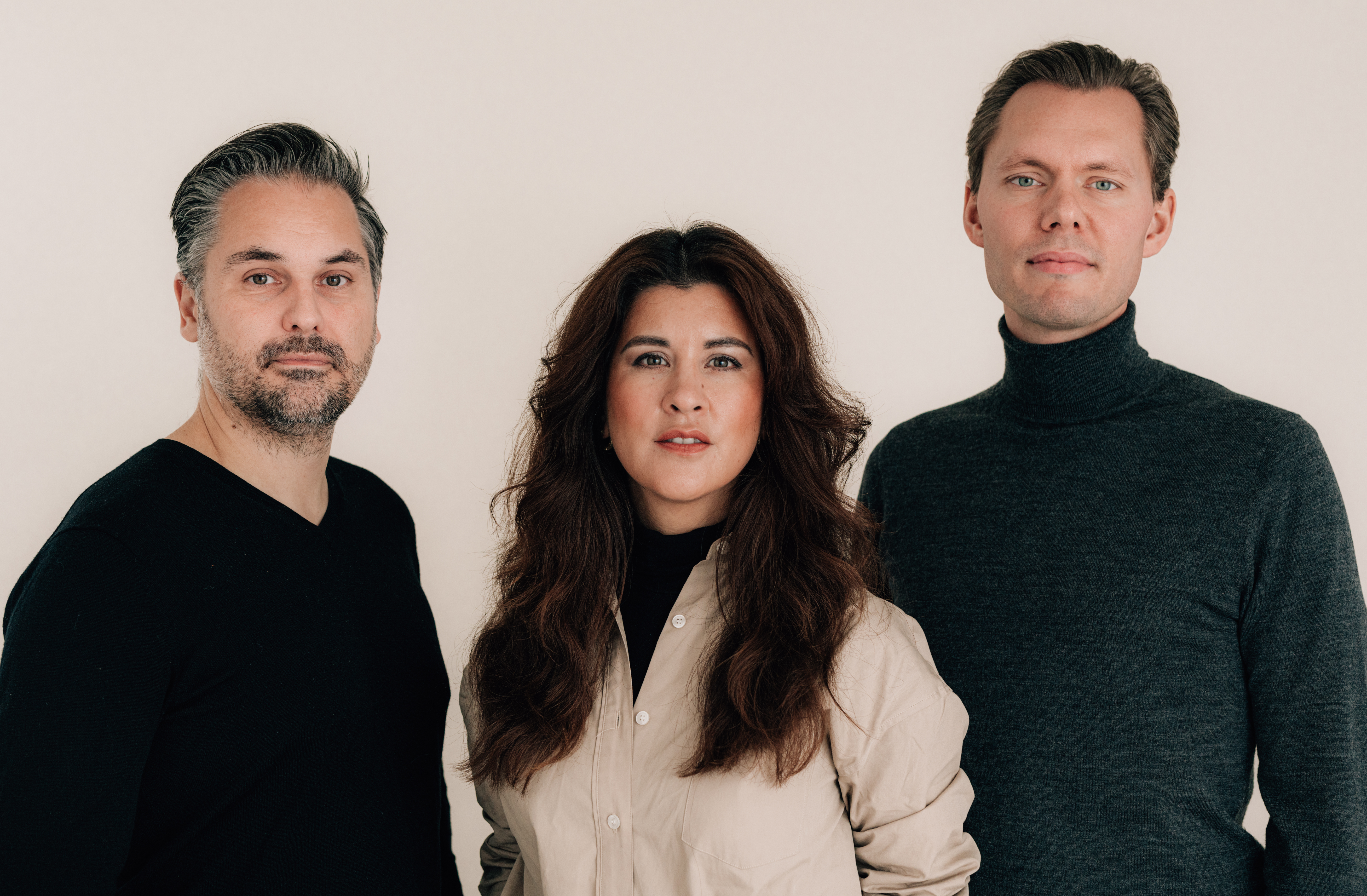 Tre personer står mot en ljus bakgrund, två män i mörka tröjor och en kvinna i beige skjorta, alla ser rakt fram utan uttryck.