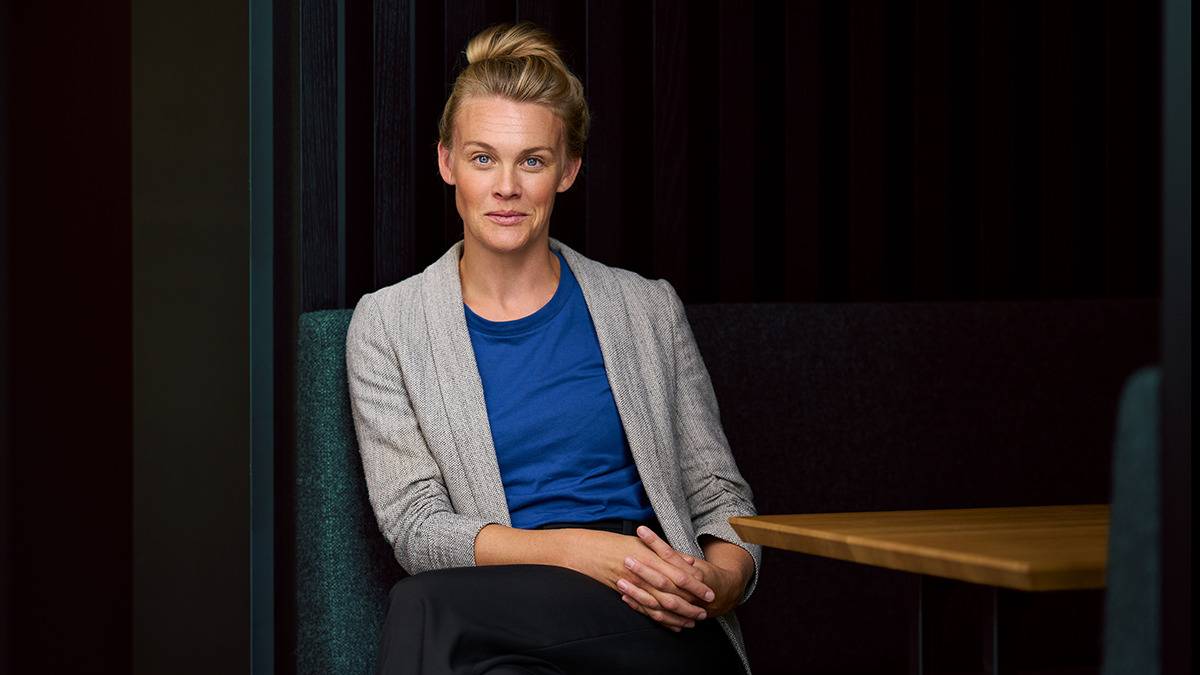 En kvinna med blont hår i knut, blå skjorta och grå kavaj sitter med händerna knäppta i en sittbås.