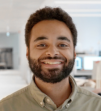 En man med kort lockigt hår och skägg ler, har på sig en ljusgrön skjorta och står i ett kontor.