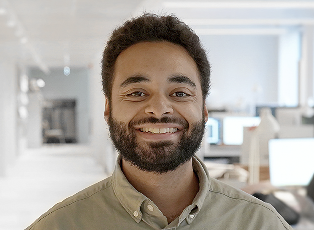En man med kort lockigt hår och skägg ler och står i ett kontor med skrivbord och datorer i bakgrunden.