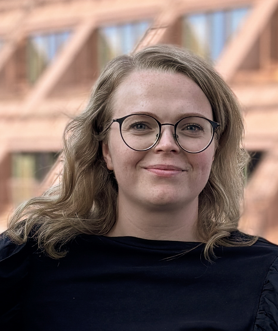 En person med långt blont vågigt hår och runda glasögon, iklädd en svart topp, står utomhus framför en geometrisk byggnad.