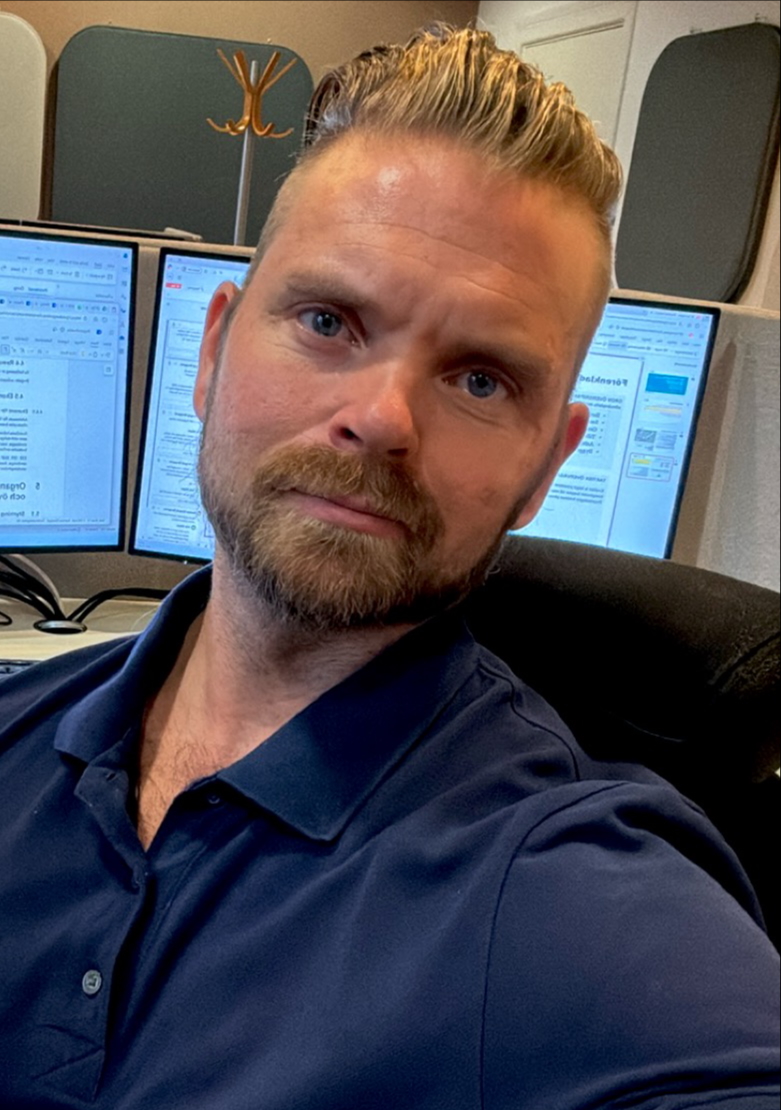 En man med skägg och kammat hår sitter i en kontorsstol, har en mörkblå pikétröja och tre datorskärmar med dokument bakom sig.