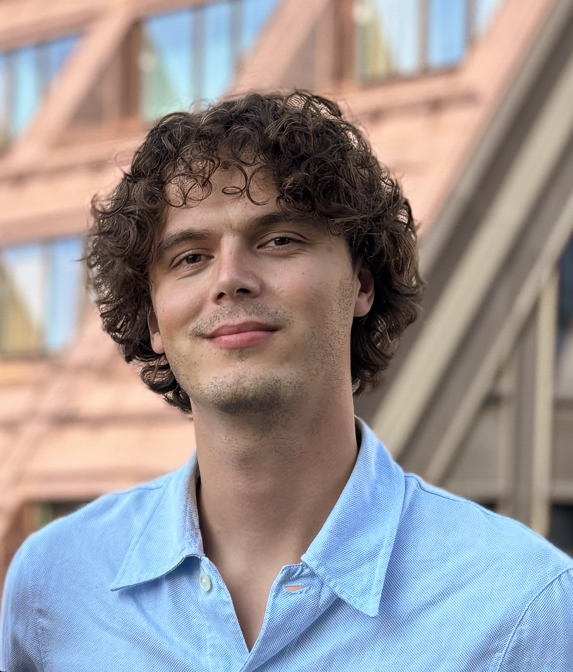 En ung man med lockigt brunt hår och ljusblå skjorta står utomhus framför en modern byggnad.