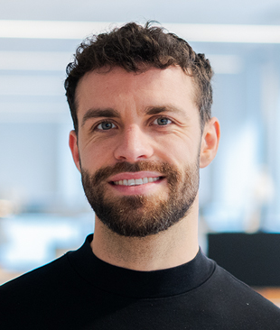 En man med kort lockigt hår och skägg, klädd i svart tröja, ler mot kameran i ett modernt kontor.