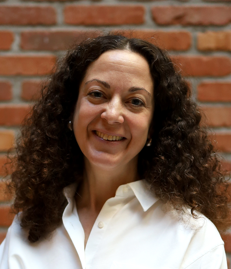 Kvinna med lockigt brunt hår som har en vit skjorta och står framför en röd tegelvägg.