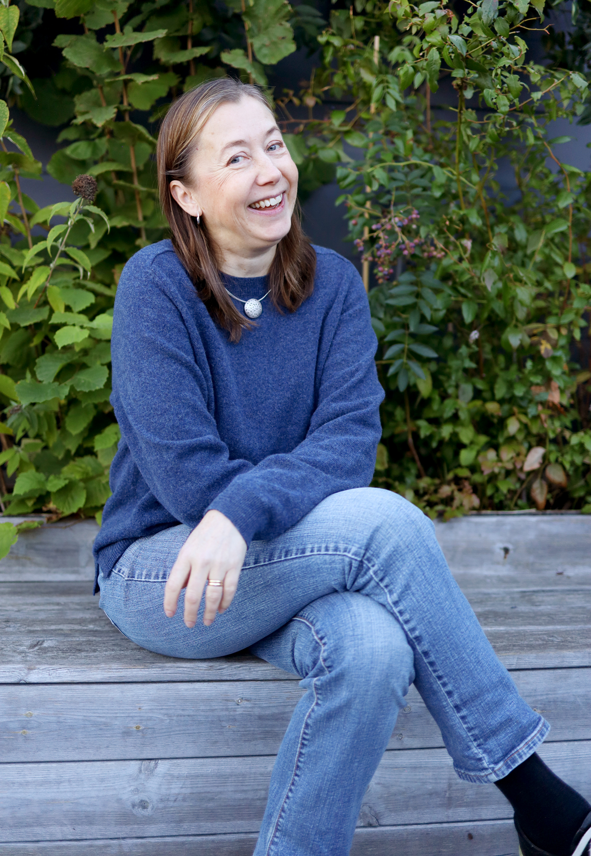 En kvinna i blå tröja och jeans sitter på en träbänk utomhus och ler, med gröna växter i bakgrunden.