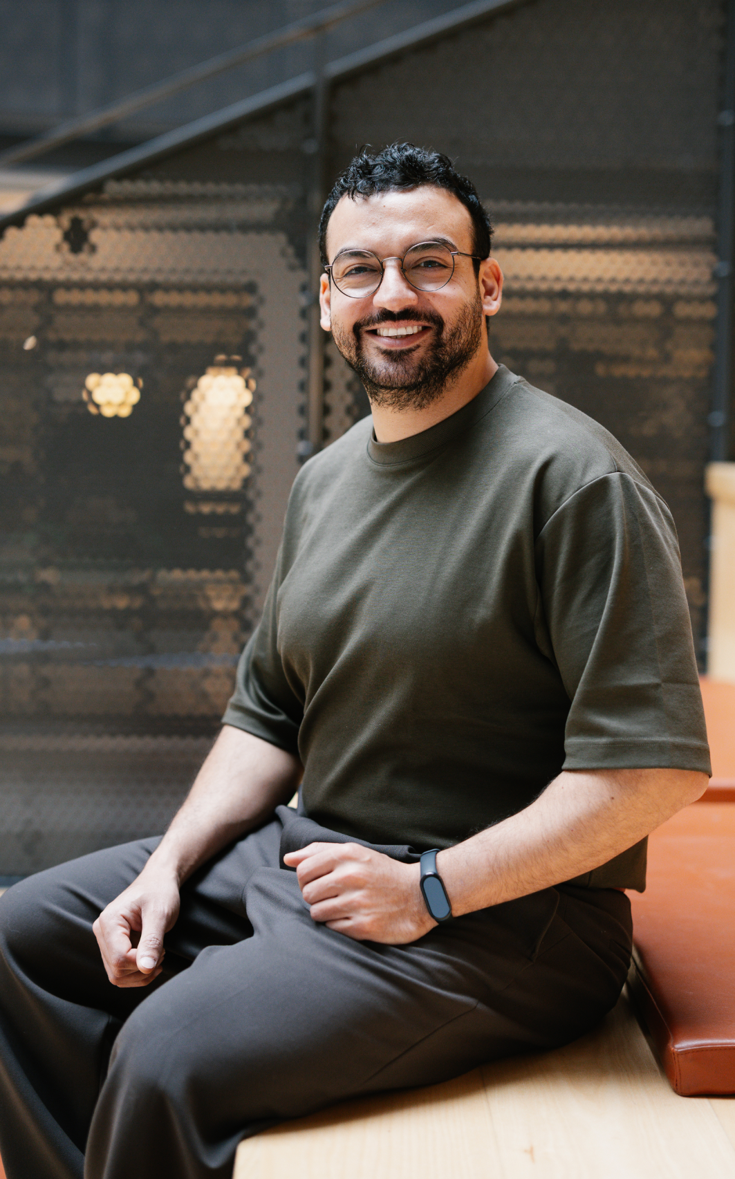 En man med glasögon och skägg, iklädd mörkgrön t-shirt och bruna byxor, sitter på en träbänk inomhus och ler mot kameran.