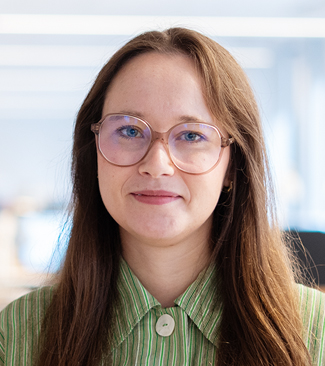 En kvinna med långt brunt hår och glasögon har på sig en grönrandig tröja och står inomhus med neutral min.