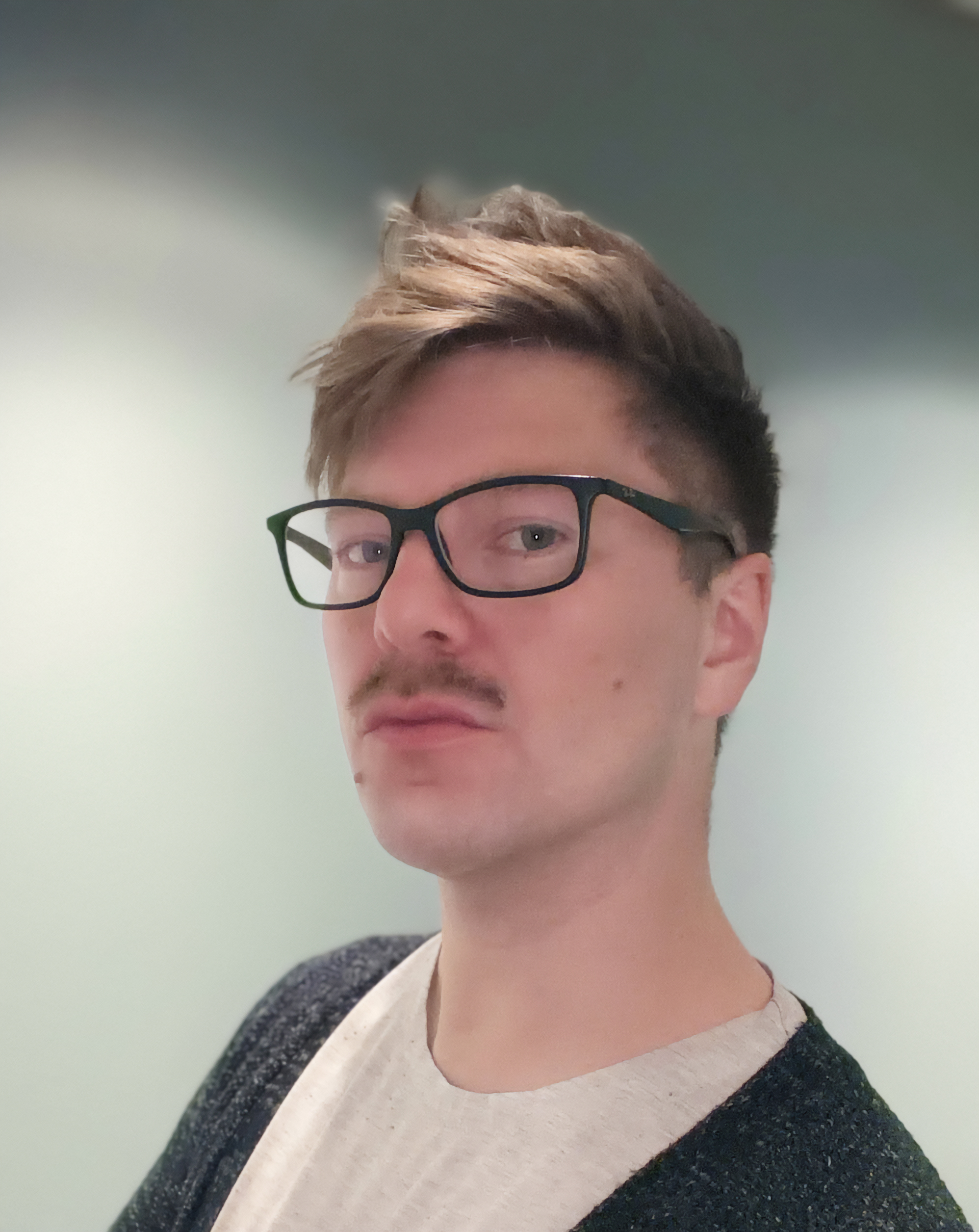 En man med kort brunt hår, glasögon och mustasch tittar mot kameran. Han har skjorta och mörk kofta mot ljus bakgrund.