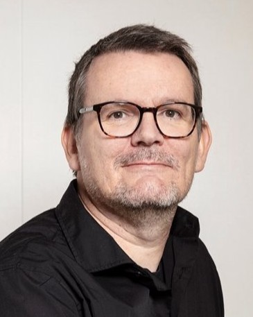 En man med kort brunt hår, glasögon och skägg, iförd svart tröja, står framför en ljus bakgrund.