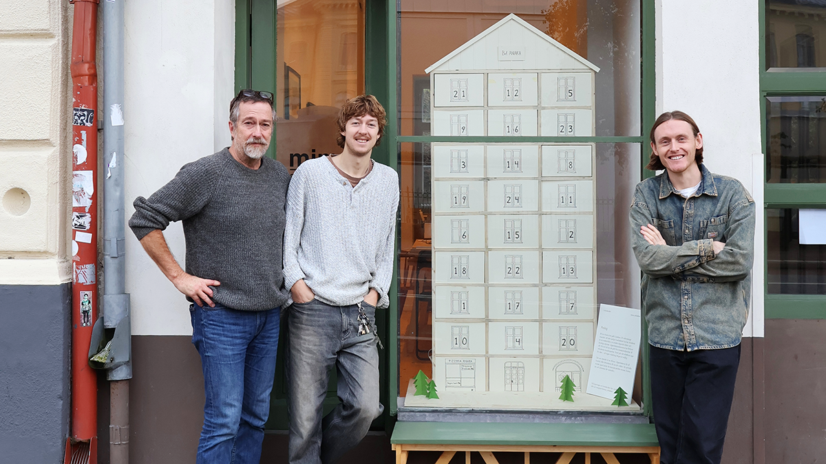 Tre personer står bredvid en handgjord modell som ser ut som en flervåningsbyggnad, placerad utanför en butik.