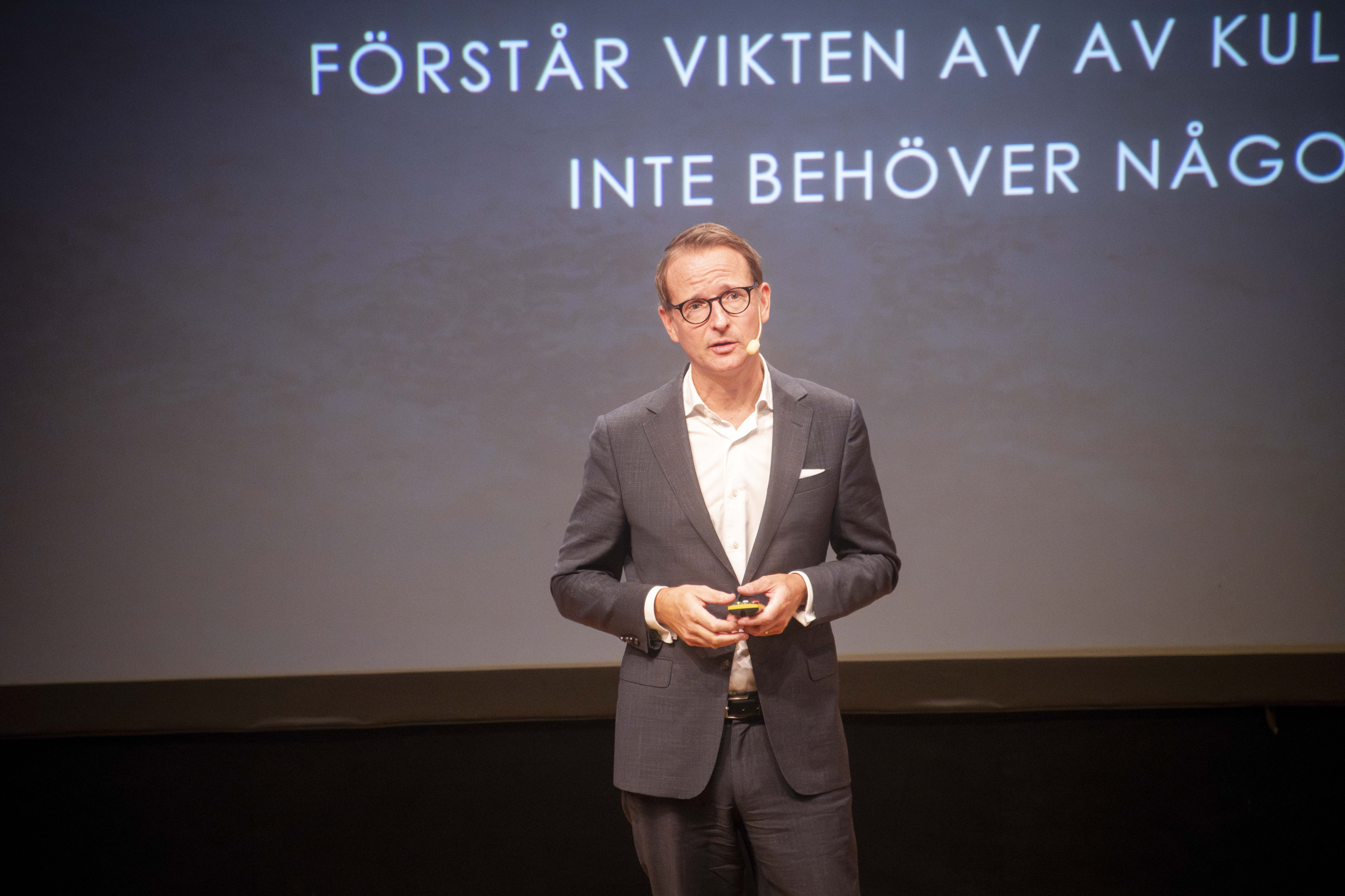 En man i kostym står på en scen och håller en presentation. Svensk text visas på en skärm bakom honom.