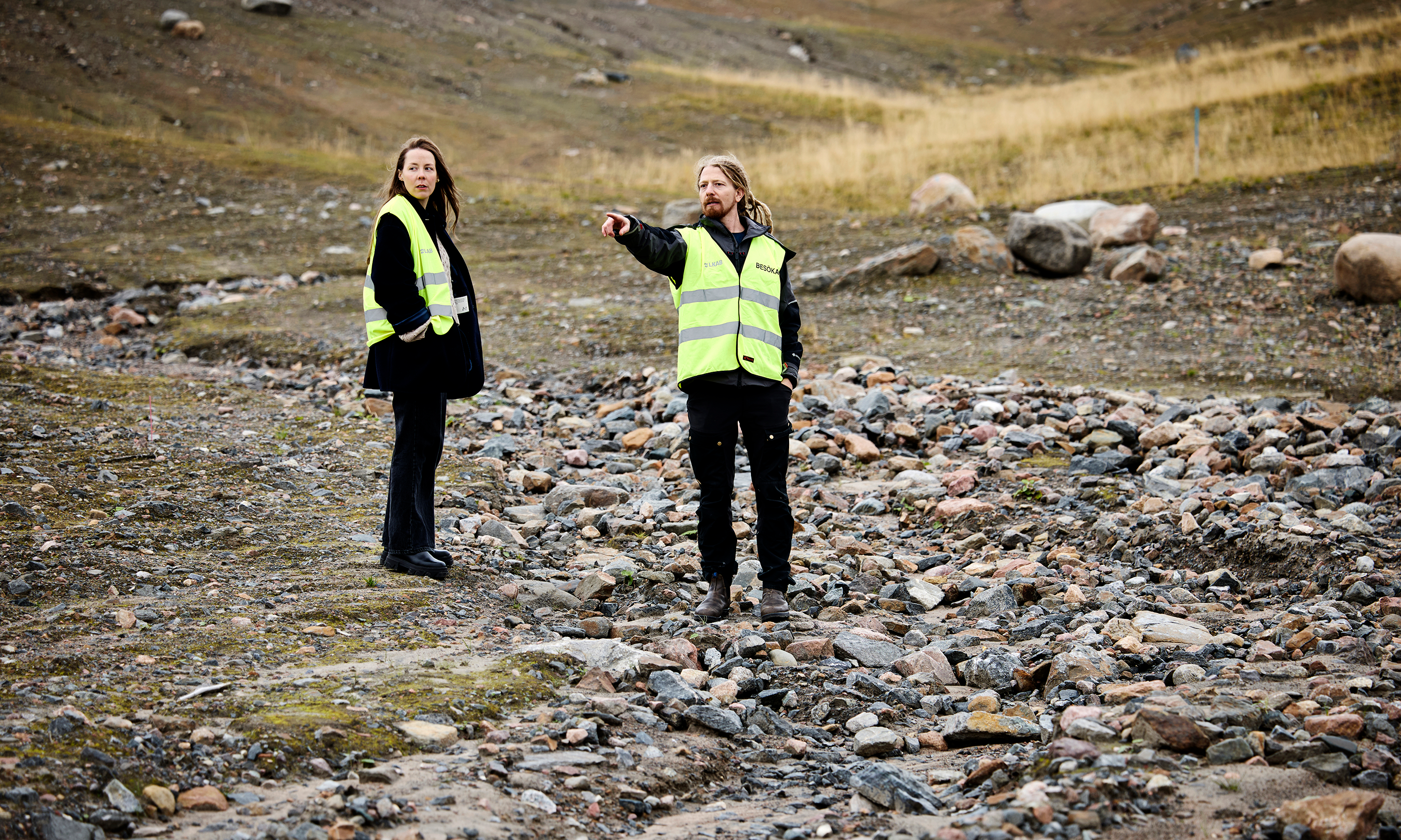 Två personer med gula säkerhetsvästar står på stenig mark; en pekar på något långt bort och den andra tittar ditåt.