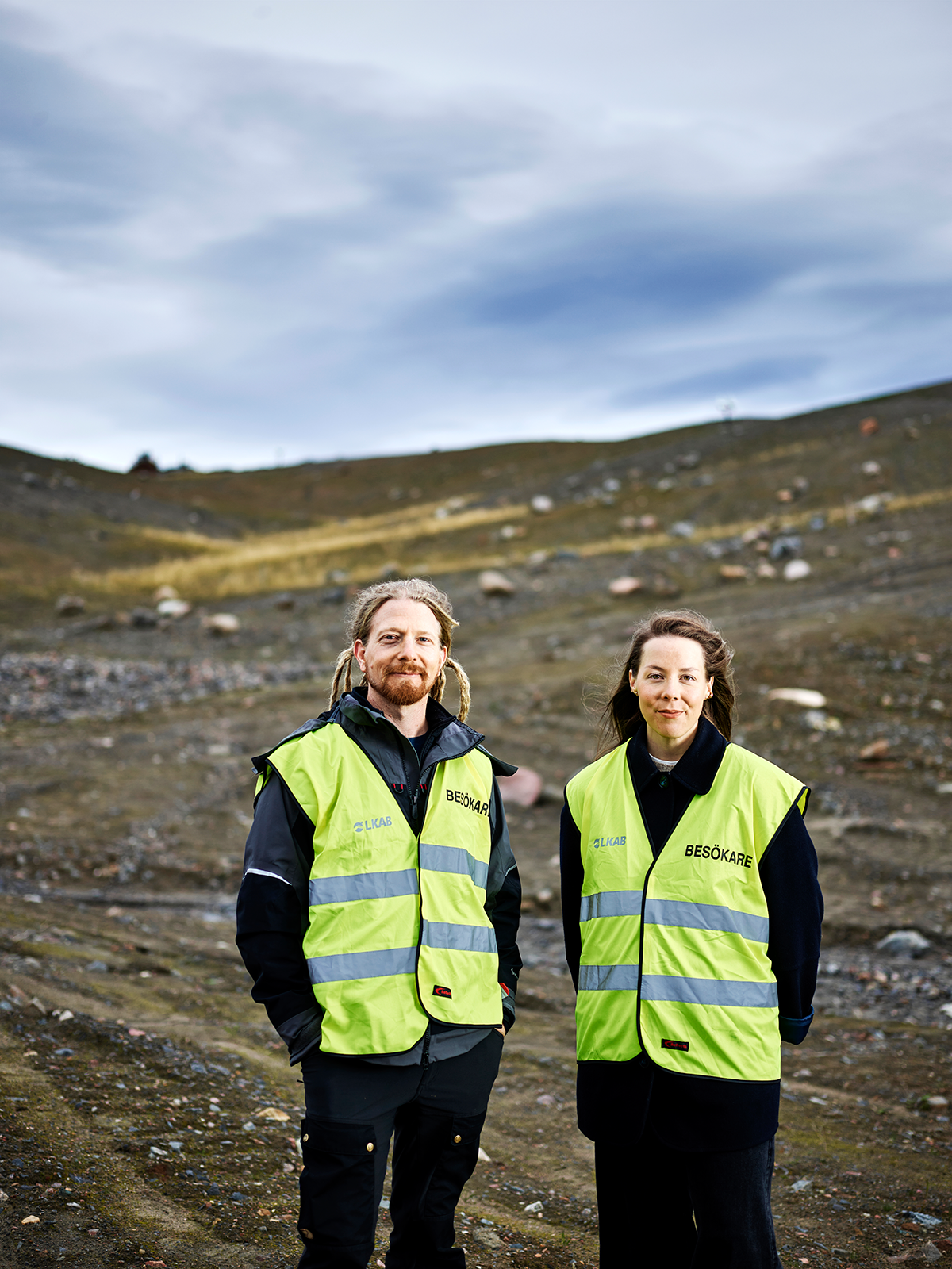 Två personer med gula reflexvästar står utomhus på stenig mark under en molnig himmel.