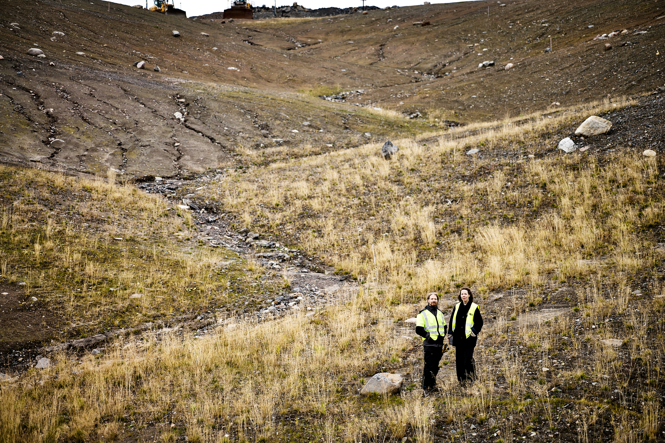 Två personer med gula säkerhetsvästar står på en sluttning med gräs, jord och stenar.