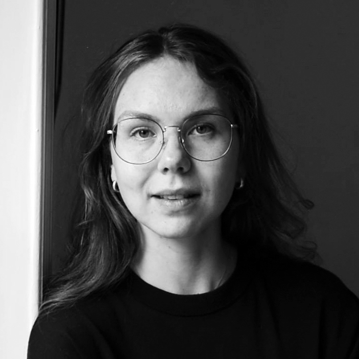 En kvinna med långt hår, glasögon och stora örhängen står vänd mot kameran i mörk tröja mot en enkel bakgrund.