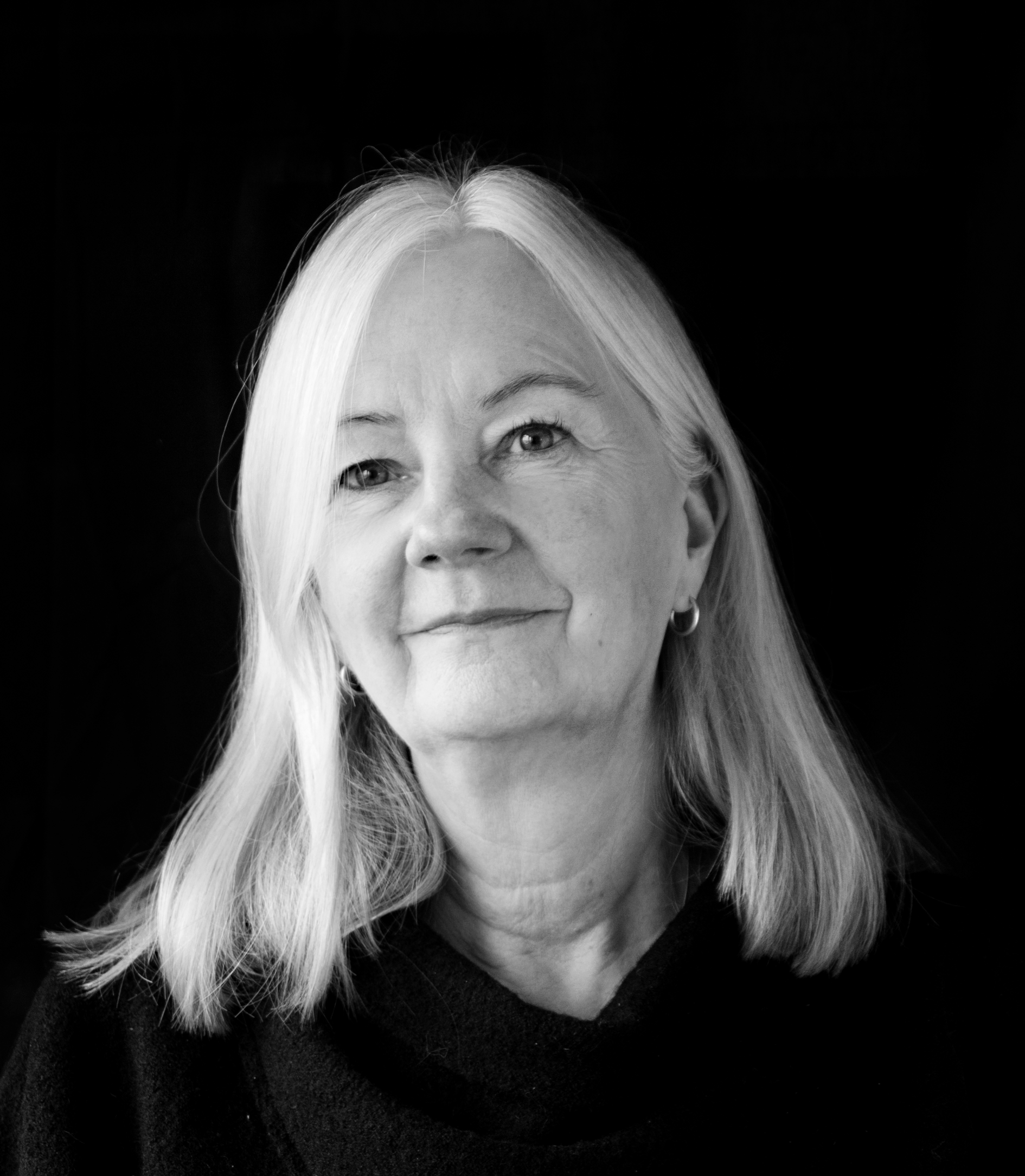 Svartvit porträttbild av en äldre kvinna med rakt axellångt hår, mörk tröja och små ringörhängen, leende mot mörk bakgrund.