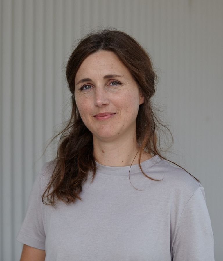 En kvinna med långt brunt hår och ljusgrå t-shirt står framför en vertikalt randig ljus bakgrund och tittar mot kameran.