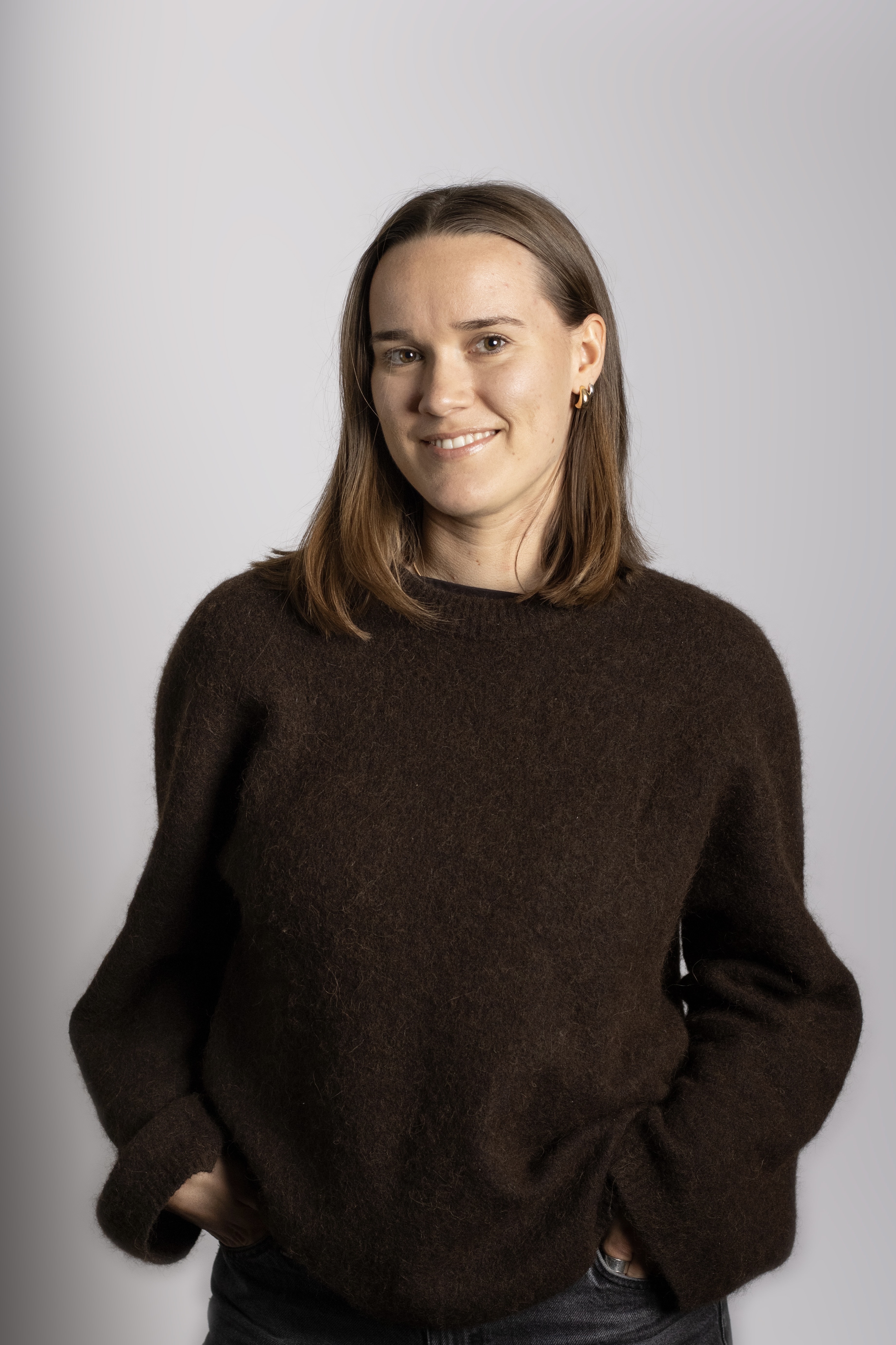 En person med rakt brunt hår har på sig en mörkbrun tröja och jeans, står mot en ljus bakgrund med händerna i fickorna och ler.