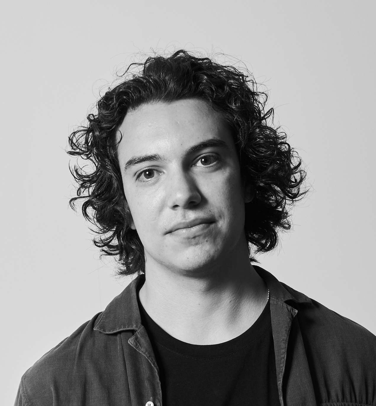 Svartvitt porträtt av en man med lockigt hår, mörk skjorta över svart t-shirt, ser in i kameran mot enkel bakgrund.