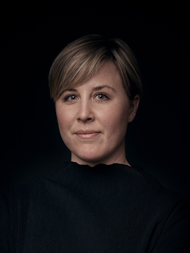 En kvinna med kort blont hår och neutral min bär en svart topp och står mot en mörk bakgrund.