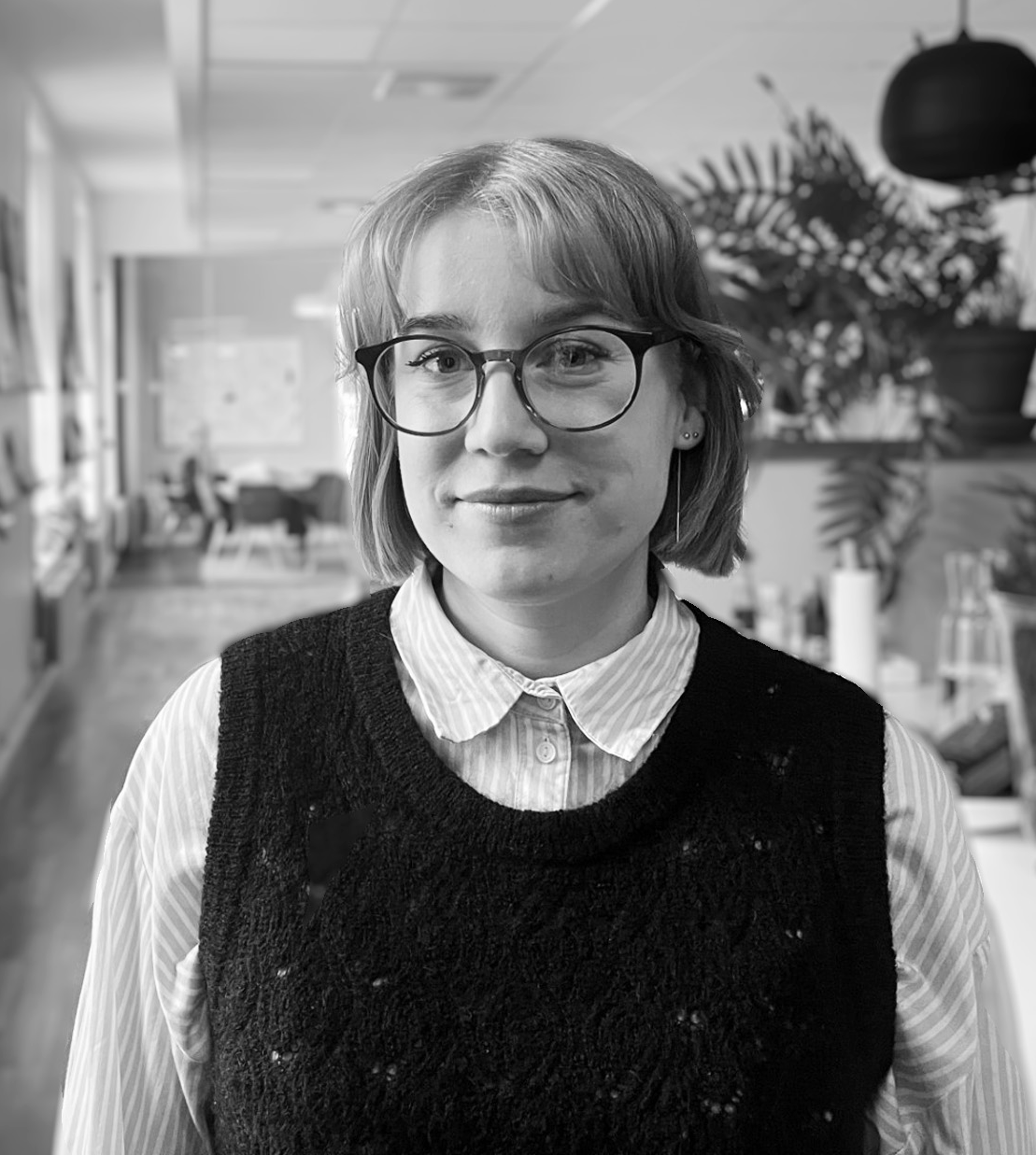 Person med glasögon och kort hår, iklädd skjorta med krage och slipover, står i ett kontor med växter och skrivbord i bakgrunden.