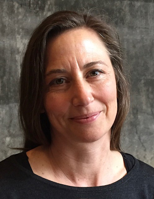 En kvinna med långt brunt hår och ett svagt leende är framför en grå bakgrund och har på sig en svart topp.