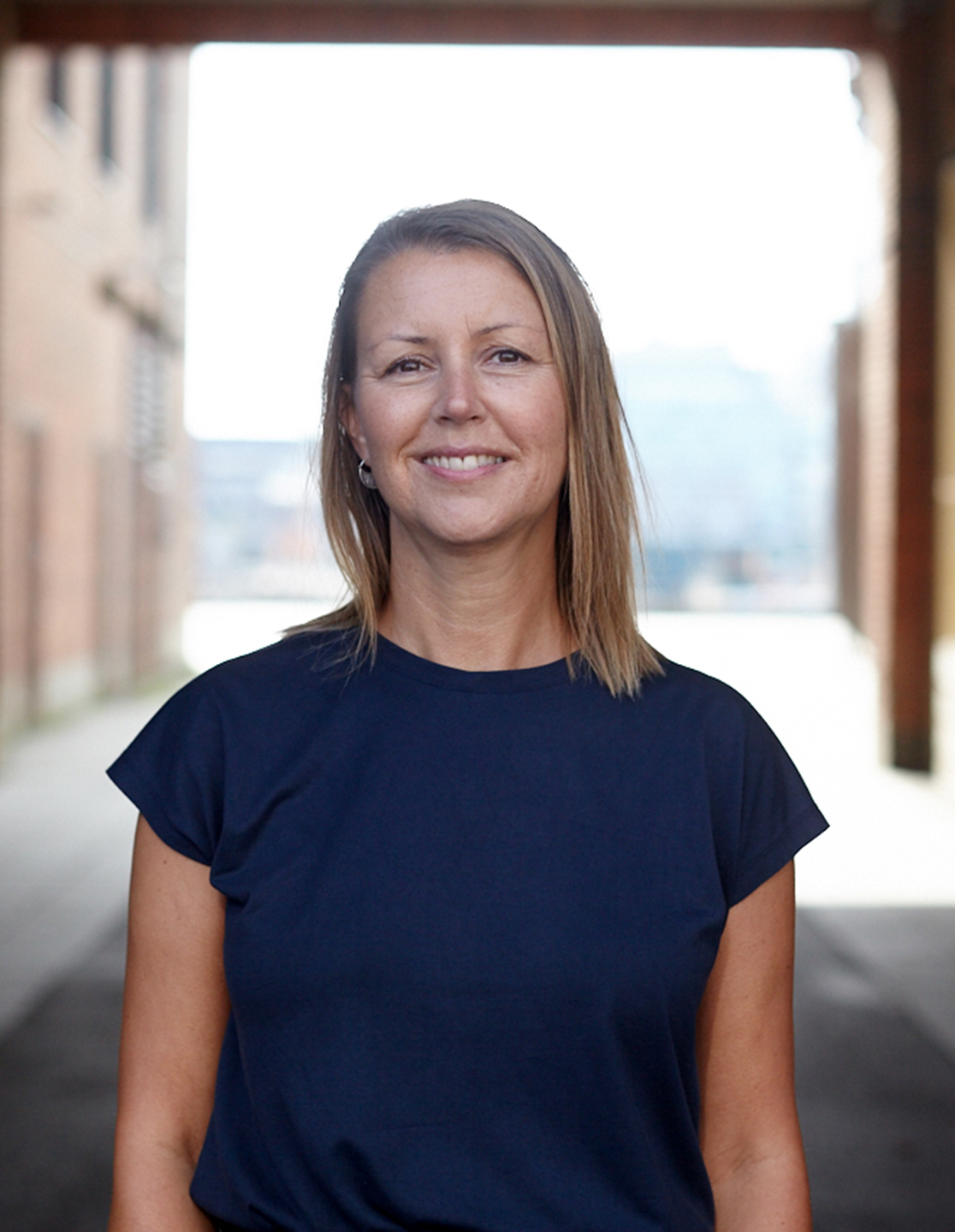 En kvinna med rakt, axellångt hår står utomhus mellan tegelbyggnader, ler mot kameran och har på sig en marinblå tröja.