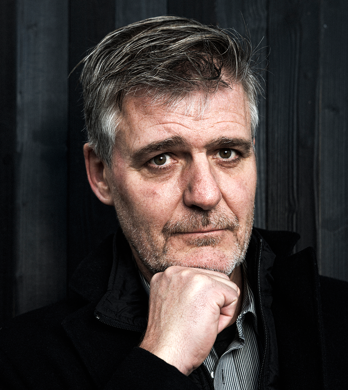 Medelålders man med grått hår och skägg tittar mot kameran, vilar hakan på handen, står framför en mörk trävägg.