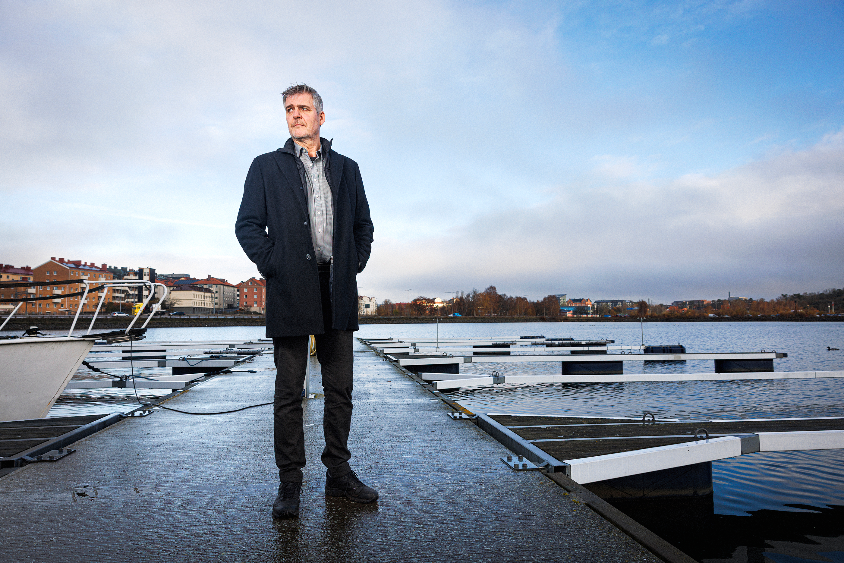 En man i mörk rock står på en våt brygga vid vattnet, med tomma båtplatser och byggnader i bakgrunden under molnig himmel.