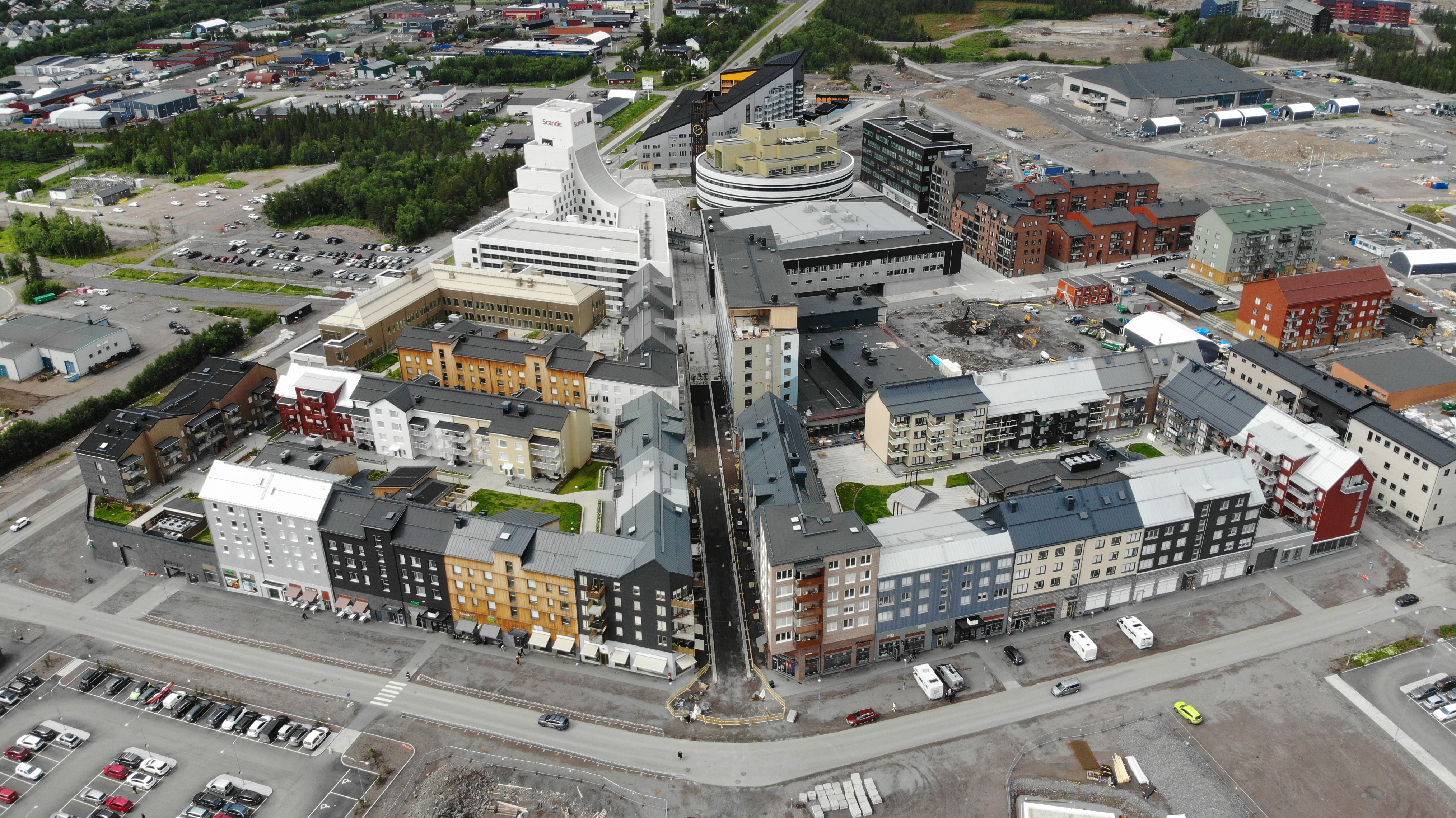 Flygfoto över ett stadsområde med byggnader längs en huvudgata, omgiven av parkeringsplatser, träd och byggarbetsplatser.